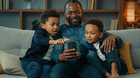 African American happy family father with ethnic sons boys little children siblings kids at home couch sofa using mobile phone together watching funny video looking at smartphone app video call chatの写真素材