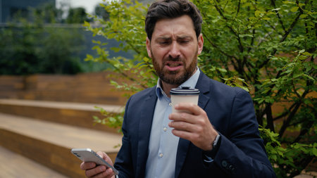 Caucasian man business employer office manager sit outside drinking coffee male guy businessman client entrepreneur using mobile phone in city outdoors bad drink disgust upset with tasteless beverageの写真素材