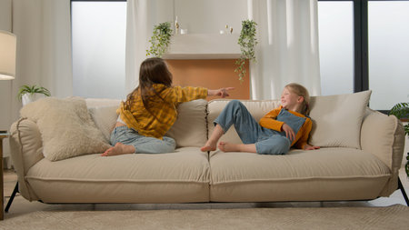 Two laughing little Caucasian children friends kids girls playing couch pointing finger gesture you sisters relationship daughters family fun talking contend debate sharing sofa together living roomの写真素材