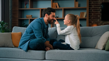 Playful caucasian young family daddy and small daughter sitting comfortable on couch loving cute girl touching father nose funny joke playing game with parent having fun spending free time togetherの写真素材