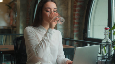 Caucasian business woman girl female businesswoman office manager worker typing laptop student distant education e-learning drinking glass of water at cafe table thirsty drink refreshment happy smileの写真素材
