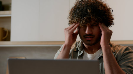 Tired Arabian male freelancer businessman work from home kitchen suffer with headache. Ill sick exhausted Indian man student guy fatigued with laptop working massage temples pain head health problemの写真素材