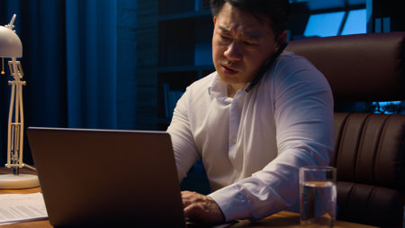 Tired angry Asian business man talking mobile phone at night evening office check documents working laptop overworked stressed Korean businessman executive manager arguing with call computer paperworkの写真素材