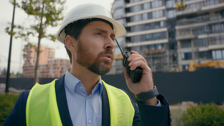 Caucasian builder architect engineer contractor repairman outdoors in city talk with remote radio building urban development industry man worker in helmet hardhat and vest speaking on walkie talkieの写真素材