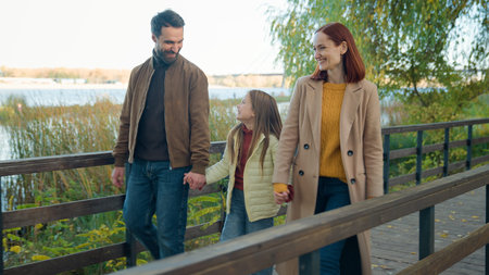 Happy Caucasian family parents couple with adopted custody adorable daughter child girl kid weekend in park autumn outdoors near lake river bridge walking talking smiling together walk holding handsの写真素材