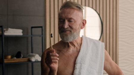 Old Caucasian retired male senior man with bath towel on shoulder showing holding bamboo tooth brush recommend brushing teeth oral care clean health mouth hygiene dental morning habit routine bathroomの写真素材