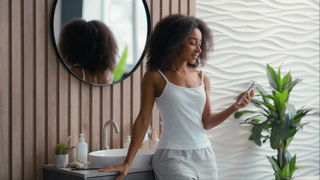 African American happy smiling woman using mobile phone browsing chatting smartphone girl telephone social media sms message shopping online internet surfing app in bathroom home beauty procedureの写真素材