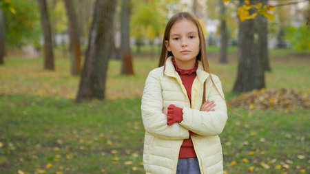 Portrait in autumn park outdoors alone sad serious confident Caucasian child girl kid daughter children schoolgirl pupil standing posing looking at camera hands crossed familyの写真素材