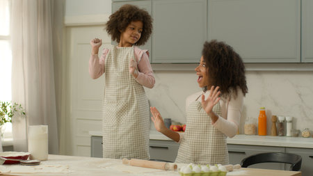 Happy African American family woman female mother mom together fooling fun at kitchen little child kid girl daughter dancing listening music dance in aprons cooking pause relax joy moving weekendの写真素材