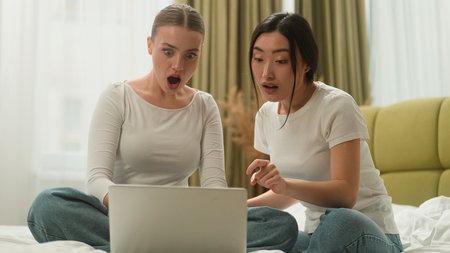 Happy smiling laughing excited multiethnic multiracial diversity two friends Caucasian Asian women girls female ladies laptop online rejoice win computer hug achieve high five yelling at bedの写真素材