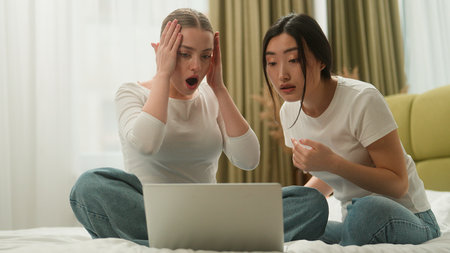 Two multiracial women upset frustrated sad worried friends female diversity girls Caucasian woman Asian girl together bed bedroom home students friendship lost problem laptop failure computer bad newsの写真素材