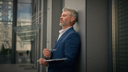 Tired exhausted Caucasian old businessman elderly employer mature business man senior male middle-aged manager with laptop finish work city outdoors take off glasses leaning to wall of office buildingの写真素材