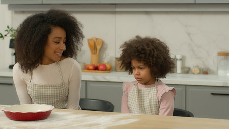 African American family custody adopted child smiling mother woman mom talking to sad upset frown shy little kid daughter offended girl at kitchen table teaching cooking baking flour cook talk problemの写真素材
