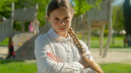 Upset sad frown little kid girl serious child daughter pupil female in city portrait schoolgirl posing crossed hands offense family worried teenager alone in park playground outdoorsの写真素材
