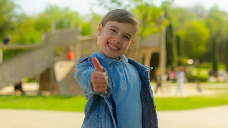 Happy little girl in city female child kid schoolgirl joyful teenager pupil student raise finger showing thumb up gesture friendly smile good sign recommend in park children playground outdoorsの写真素材