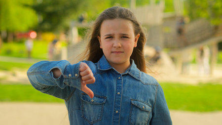 Sad upset displeased little girl schoolgirl child serious kid daughter in city outdoors looking camera showing thumb down finger gesture dislike not low mark disapprove sign childhood custody problemの写真素材