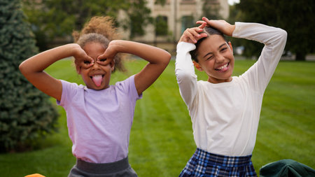 Happy little girls two kids schoolgirls children school education students pupils classmates multiracial African Indian female friends laugh have fun eyes circles grimace play game kidding outdoorsの写真素材
