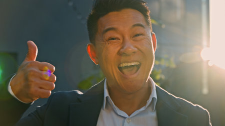 Portrait happy smiling toothy satisfied successful glad Asian 40s middle-aged man boss leader businessman standing outdoors in sun lights in sunbeams showing thumb up agree good gesture looking cameraの写真素材