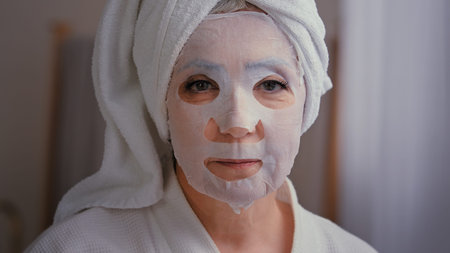 Old senior mature Caucasian woman apply skincare textile skin mask on female face showing ok good beauty cosmetology procedure in bathroom 60s lady wrap towel on head put moisturizing lifting cosmeticの写真素材