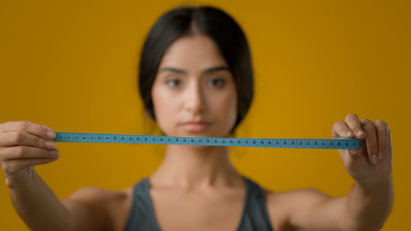 Sad upset worried Indian woman girl in yellow studio with measuring tape dissatisfied with body measure unhappy weight size shape fitness result waist fat diet unhealthy depressionの写真素材