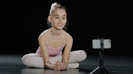 Caucasian girl child teen ballerina gymnast sitting on floor in dance hall waving hello to mobile...