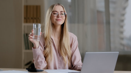 Caucasian busy business woman worker girl female student wearing glasses with laptop paperwork learning feeling thirsty drinking from glass water balance health care diet refreshment daily good habitの写真素材