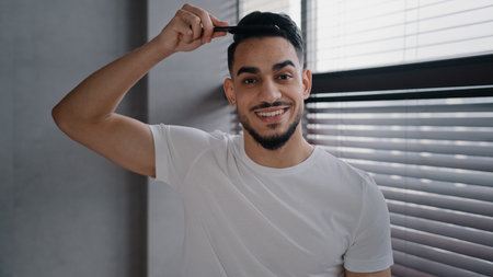Arabian spaniard arab man looking at camera combing hair with hairbrush getting ready to date work day home guy in white T-shirt morning beauty routine styling haircut stylish preparing for datingの写真素材