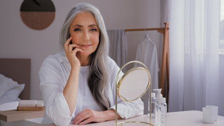 Caucasian old beautiful 60s senior lady mature aging elderly model woman with gray hair female granny grandmother looking at camera sitting at home with mirror smile wrinkled face skin care procedureの写真素材