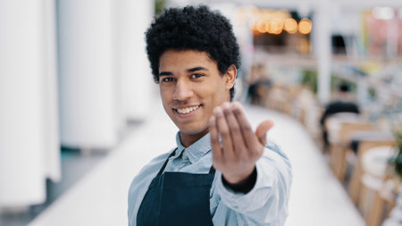 Friendly african american young guy male waiter salesman restaurant cafe kitchen bar worker in apron looking at camera showing gesture her you come here approach welcome inviting customers promotionsの写真素材