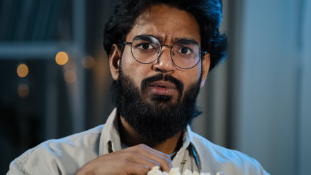 Male home portrait serious interested attentive Arabic Hispanic Indian bearded man guy in glasses eating popcorn watching TV show series late evening night watch movie online sport game championshipの写真素材