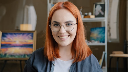 Close-up happy woman with red hair painter girl artist in glasses looks at camera stretches forward hands smeared in acrylic paint laughs inspired by drawing decor design artの写真素材