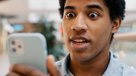 African happy excited guy surprised by message good news winning online bets looking at mobile phone has offer invite opens mouth in surprise delighted shock man feels amazing wonder with achievementの写真素材