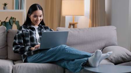 Carefree Asian girl enters living room sit down on comfortable sofa takes laptop with smile and watches video online woman uses computer sitting on couch at home casual browsing net resting typingの写真素材