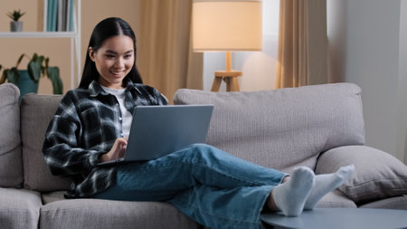 Smiling asian woman using laptop casual browsing searching online play computer game. Korean student girl freelancer sitting on couch e-learning internet education chatting studying working with wi-fiの写真素材