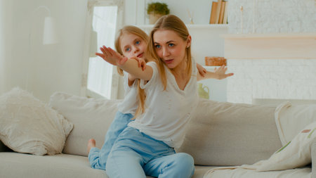 Caucasian mother with little daughter piggyback pretending flying stretch hands to side playing airplane game sitting at couch in living room. Mom with child play kid girl having fun at sofa at homeの写真素材