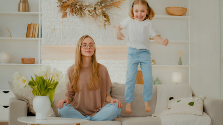 Mindful calm Caucasian mother in glasses meditating sitting on sofa while active child daughter jumping on couch. Peaceful mom relaxing in yoga meditation at home little kid girl jumpの写真素材