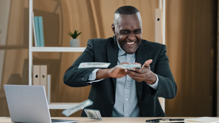 Happy successful old African American businessman senior mature man worker investor in office sitting at table holding fan of money throwing dollars banknotes enjoying winning financial prize awardの写真素材