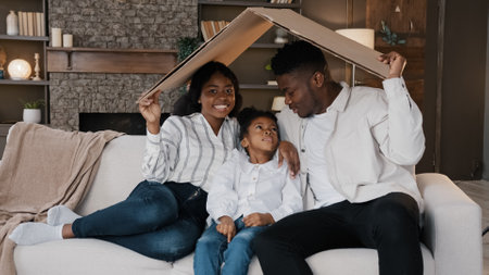 African american family parents with daughter little girl adopted child sit on sofa under cardboard roof dreaming about own house relocation home insurance real estate buying flat kid protectionの写真素材