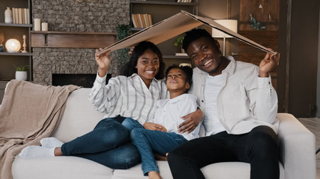 African couple sitting on couch under cardboard roof smiling looking at camera active happy daughter girl running sits on sofa. Relocation day to new home moving to own apartment homeowner portraitの写真素材