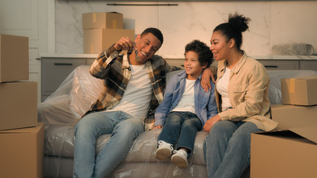 Happy family African American parents little son boy together on couch moving day relocation father holding keys of new own house buy rent apartment surprise mother child kid rejoice credit mortgageの写真素材