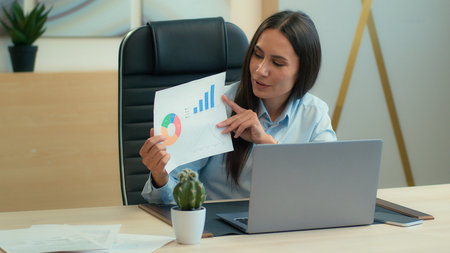 Cheerful happy Caucasian woman smiling business girl female businesswoman expert manager discuss talk video call online on laptop in office showing papers diagram sales charts graphs data analyzingの写真素材