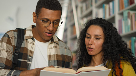 Two students African American man Arabian Latino woman together reading book textbook library...