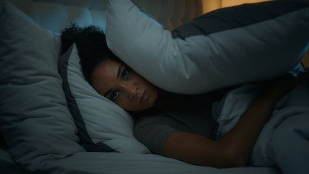 Noisy neighbours problem annoyed African American woman lying in bed night dark bedroom suffer from sleeping disorder insomnia sleepless angry girl trying sleep awake to noise cover ears with pillowsの写真素材
