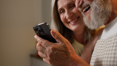 Happy mature couple buy products online retired old middle-aged adult bonding family in kitchen. Caucasian elderly man woman look in smartphone talking laughing order delivery food with mobile phoneの写真素材