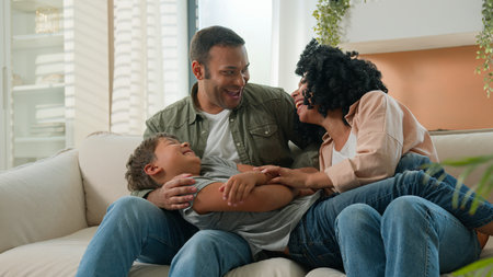 Happy multiracial family parents playing with cute little boy son tickling have fun laugh together funny game on home sofa playful African American mom dad embrace child kid affectionate relationshipの写真素材