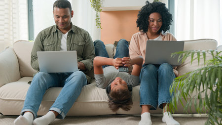 Addicted family use gadgets devices little kid child son boy lying upside down on sofa play video game on mobile phone smiling African American parents working laptops different gen using modern techの写真素材
