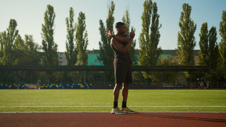 Healthy active sportsman training morning workout on city stadium stretch hands stretching shoulders muscles warm-up exercise male athlete African American fitness athletic man doing sport outdoorsの写真素材