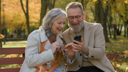 Caucasian retired couple family in love mature woman man sit on bench autumn park using mobile phone gadget together smiling wife husband spouses reading news old age and smartphone in city outdoorsの写真素材