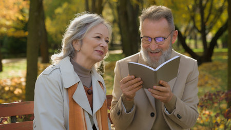 Retired Caucasian couple gray-haired grandparents reading book pension leisure time in autumn park happy old 70s aged loving man husband and wife woman relaxing on bench together retirement outdoorsの写真素材