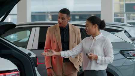 African American woman dealer seller help man client in auto dealership store saleswoman consult showing automatic trunk opening function of electric car modern automobile features shocked buyer smileの写真素材